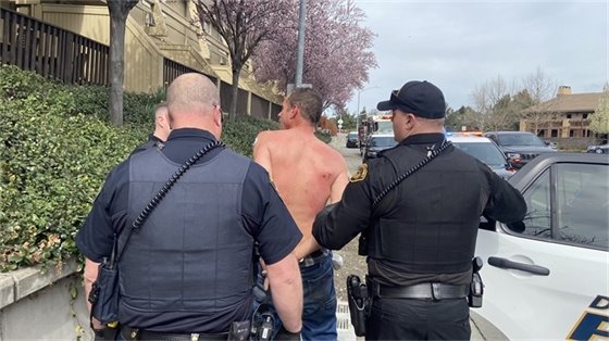 Two police officers wearing uniforms and bullet proof vests escort a handcuffed, shirtless man, to the open door of a police squad car.   