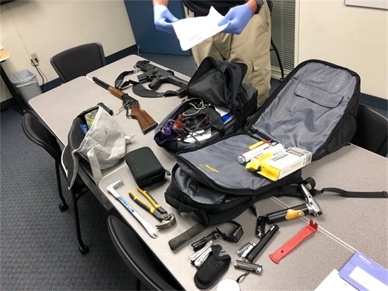 Table covered in stolen items. Three fake weapons are displayed, two open backpacks with a variety of items in them, and some electronic gadgets are on the table.  